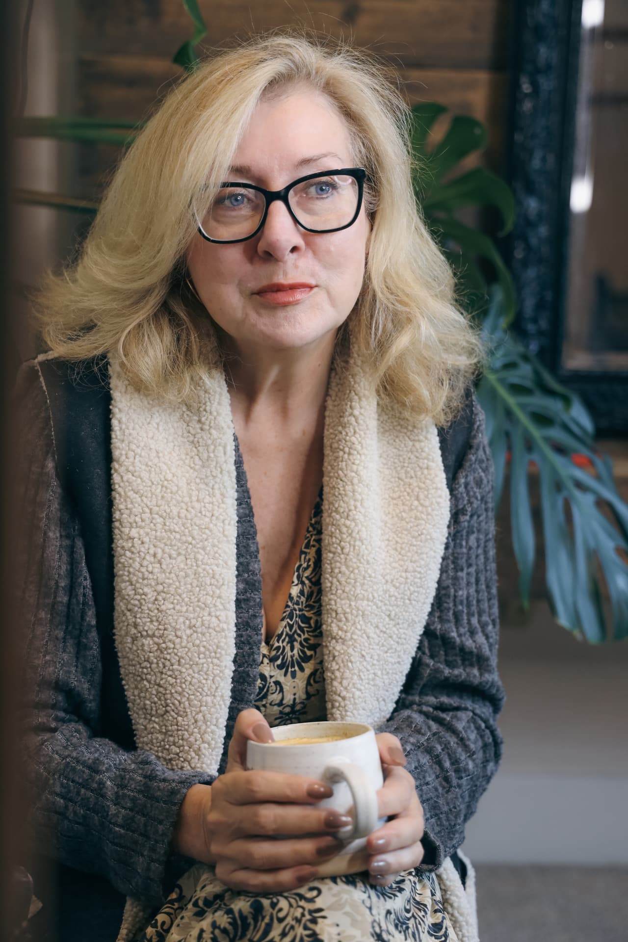 Sharon seated indoors, wearing glasses and holding a cup