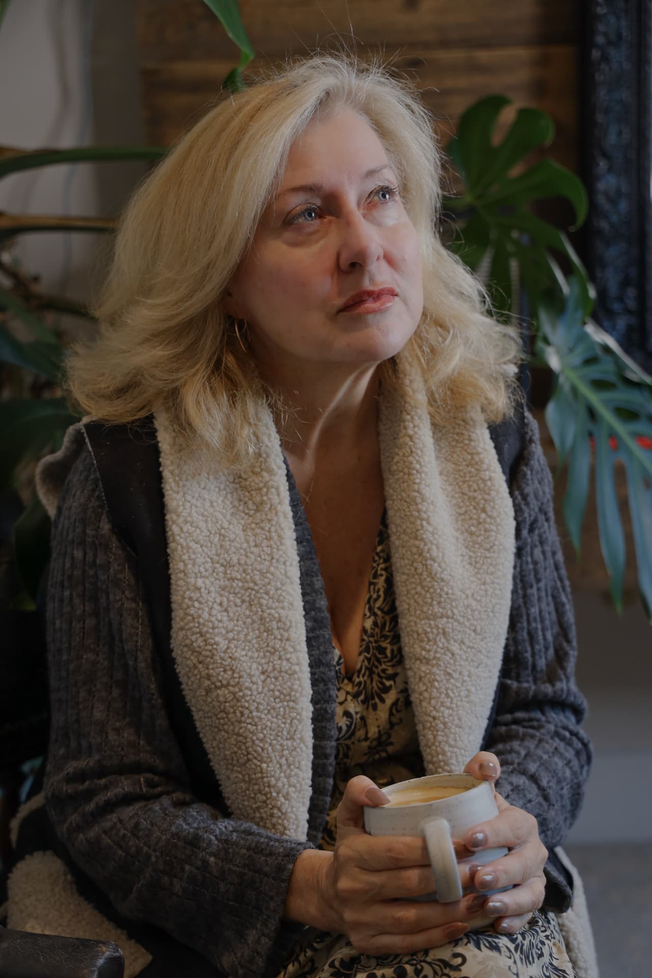 Sharon seated indoors holding a cup, looking thoughtfully away